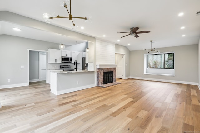 unfurnished living room with sink, a brick fireplace, vaulted ceiling, ceiling fan with notable chandelier, and light wood-type flooring