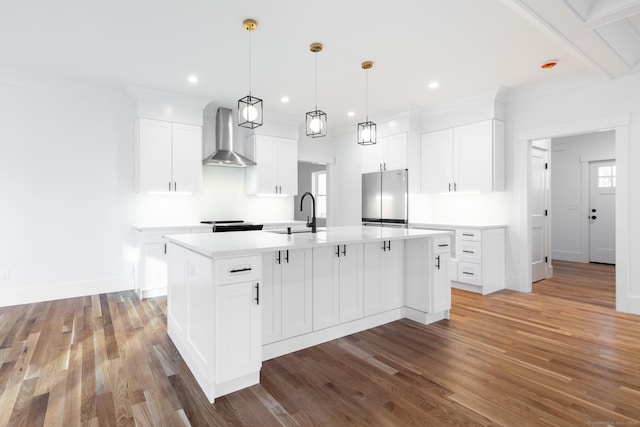 kitchen featuring white cabinets, hanging light fixtures, wall chimney exhaust hood, stainless steel fridge, and an island with sink