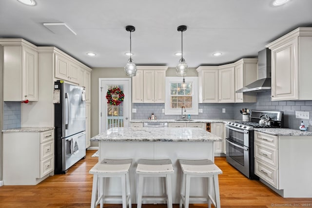 kitchen featuring wall chimney range hood, hanging light fixtures, light wood-type flooring, appliances with stainless steel finishes, and a kitchen island