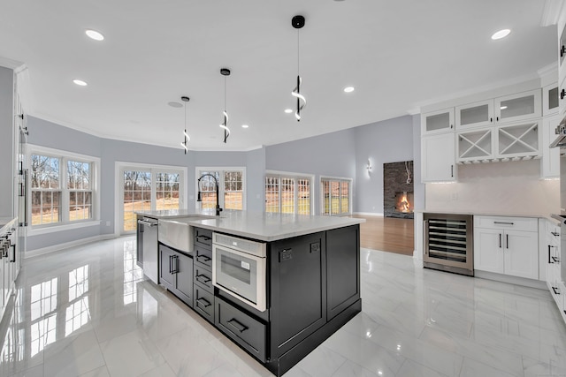 kitchen featuring beverage cooler, decorative light fixtures, a kitchen island with sink, a fireplace, and white cabinets