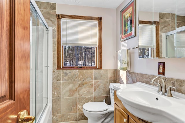 full bathroom with combined bath / shower with glass door, vanity, toilet, and tile walls