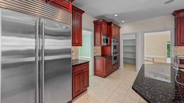 kitchen featuring light tile patterned floors, built in appliances, dark stone countertops, and backsplash