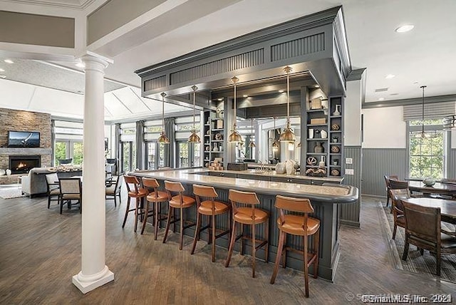 bar with dark wood-type flooring, crown molding, ornate columns, a fireplace, and decorative light fixtures