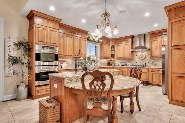 kitchen with light stone countertops, appliances with stainless steel finishes, wall chimney range hood, a chandelier, and an island with sink