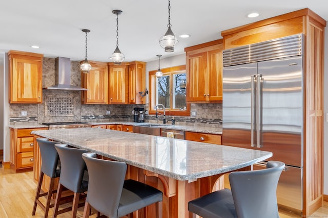 kitchen featuring wall chimney exhaust hood, a kitchen island, pendant lighting, stainless steel appliances, and light stone countertops