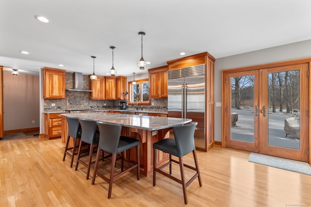 kitchen with built in fridge, decorative light fixtures, a center island, light stone countertops, and wall chimney range hood