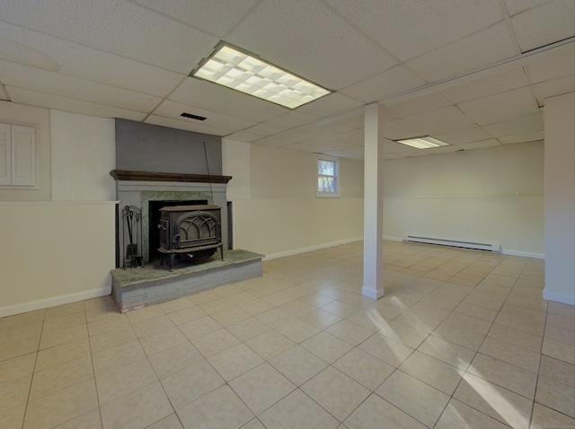 basement featuring a drop ceiling, a wood stove, light tile patterned floors, and a baseboard heating unit