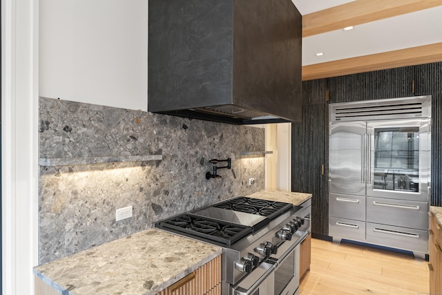 kitchen with high quality appliances, wall chimney exhaust hood, light wood-type flooring, tasteful backsplash, and light stone counters