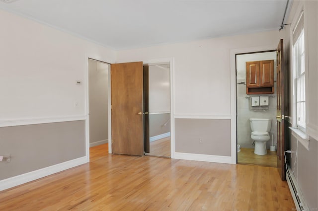 unfurnished bedroom featuring connected bathroom, baseboard heating, crown molding, light hardwood / wood-style floors, and a closet