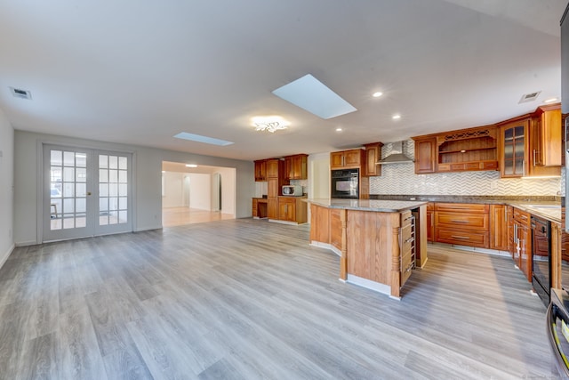 kitchen featuring a skylight, wall chimney range hood, light hardwood / wood-style flooring, black oven, and a kitchen island