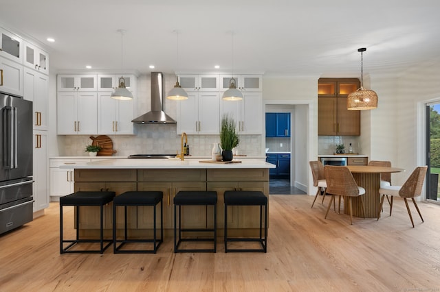 kitchen featuring high end fridge, wall chimney range hood, pendant lighting, a center island with sink, and white cabinets