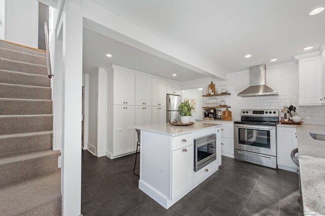 kitchen featuring light stone countertops, wall chimney exhaust hood, stainless steel appliances, a kitchen island, and white cabinetry