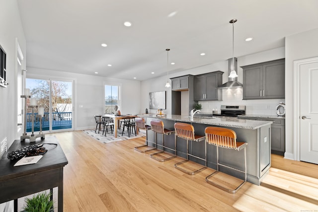 kitchen with wall chimney range hood, light hardwood / wood-style flooring, stainless steel range oven, decorative light fixtures, and a center island with sink