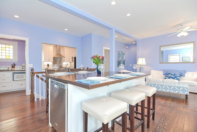 kitchen featuring light brown cabinetry, wall chimney exhaust hood, dishwasher, dark hardwood / wood-style floors, and a breakfast bar area