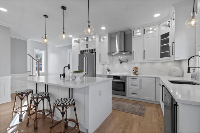 kitchen with a kitchen island with sink, wall chimney range hood, hanging light fixtures, appliances with stainless steel finishes, and white cabinetry