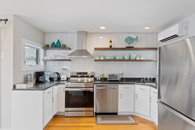 kitchen with a wall mounted air conditioner, appliances with stainless steel finishes, wall chimney exhaust hood, sink, and white cabinets