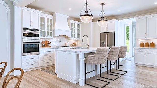 kitchen featuring premium range hood, white cabinetry, hanging light fixtures, stainless steel appliances, and an island with sink