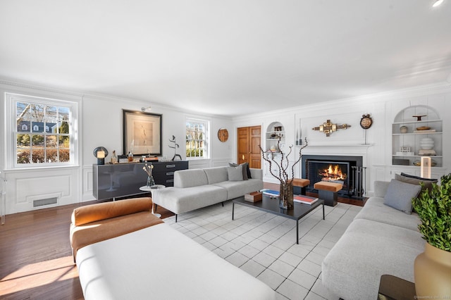 living room with a warm lit fireplace, visible vents, built in features, ornamental molding, and a decorative wall