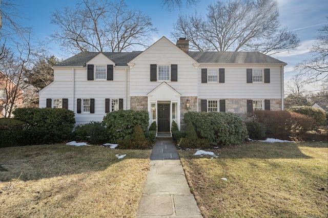 view of front facade featuring stone siding, a chimney, a front yard, and roof with shingles