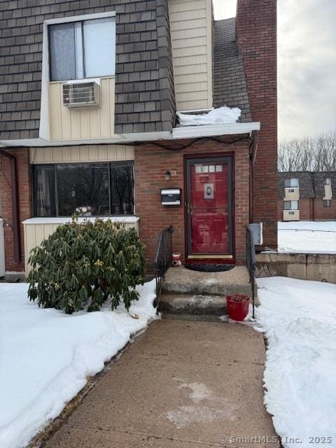 snow covered property entrance with cooling unit, roof with shingles, brick siding, and a chimney