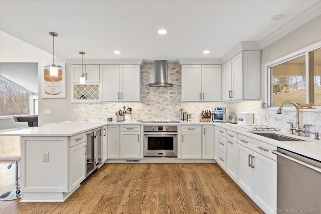 kitchen featuring wall chimney range hood, sink, appliances with stainless steel finishes, decorative light fixtures, and white cabinetry