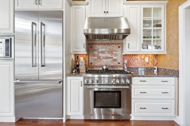 kitchen with built in appliances, white cabinets, exhaust hood, and dark stone counters