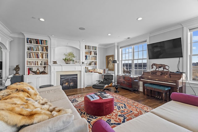 living room with dark wood-style floors, built in shelves, crown molding, recessed lighting, and a glass covered fireplace