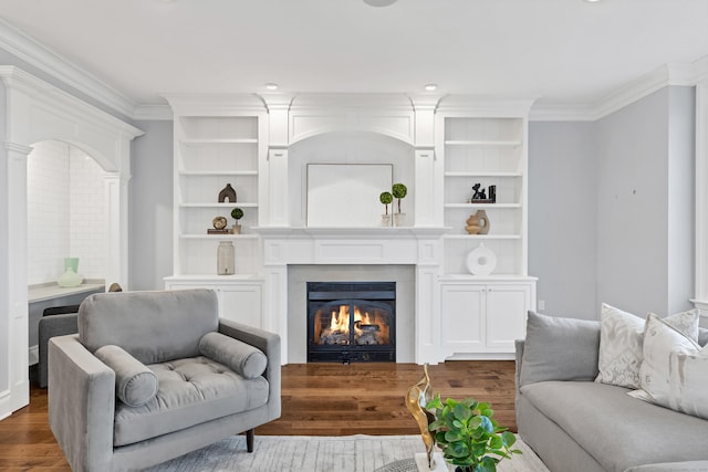 living room featuring built in shelves, crown molding, a warm lit fireplace, and dark wood finished floors
