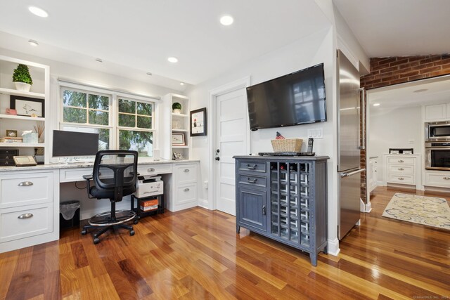 home office with light hardwood / wood-style flooring, built in desk, lofted ceiling, and brick wall