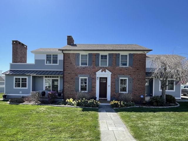 colonial-style house with a chimney, metal roof, a standing seam roof, a front yard, and brick siding