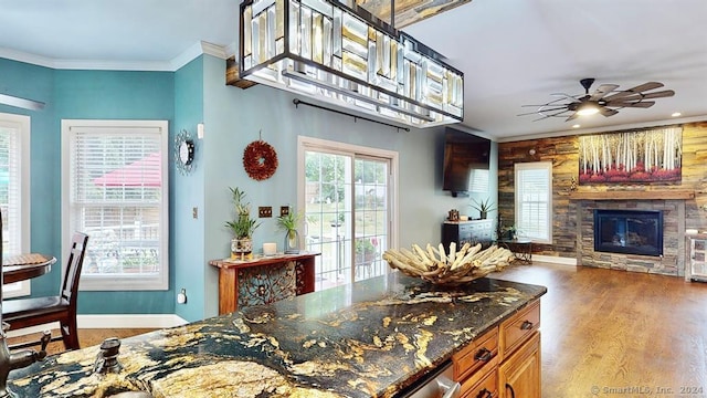 kitchen featuring ornamental molding, dark stone counters, ceiling fan, light hardwood / wood-style flooring, and a stone fireplace