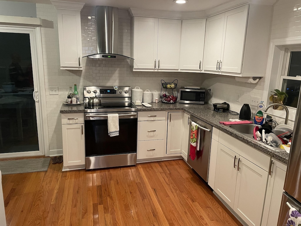 kitchen with sink, light hardwood / wood-style flooring, wall chimney exhaust hood, appliances with stainless steel finishes, and white cabinetry