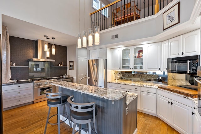 kitchen with white cabinets, a center island with sink, high end appliances, and wall chimney range hood