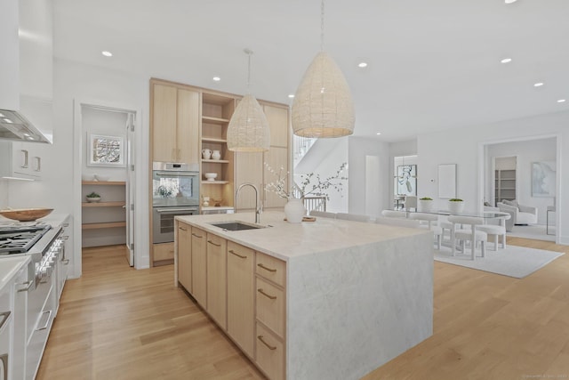 kitchen with sink, wall chimney exhaust hood, stainless steel double oven, hanging light fixtures, and a center island with sink