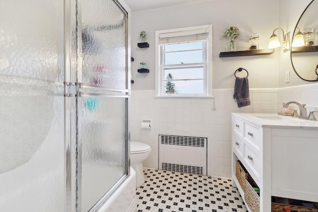 full bathroom featuring vanity, bath / shower combo with glass door, toilet, tile walls, and radiator heating unit