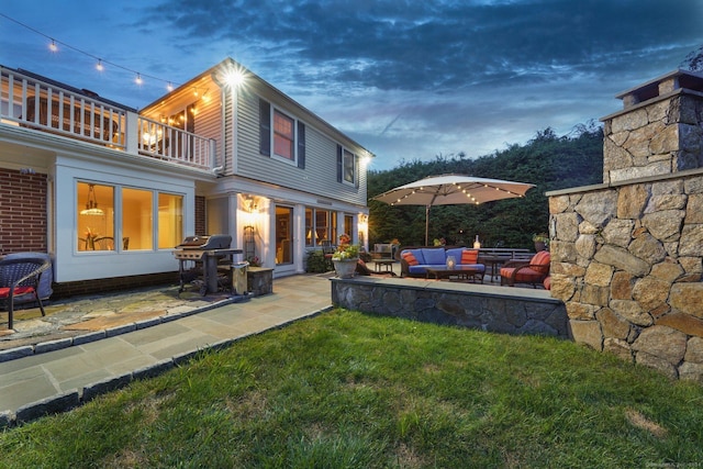 back house at dusk with an outdoor living space with a fireplace, a balcony, a patio, and a lawn