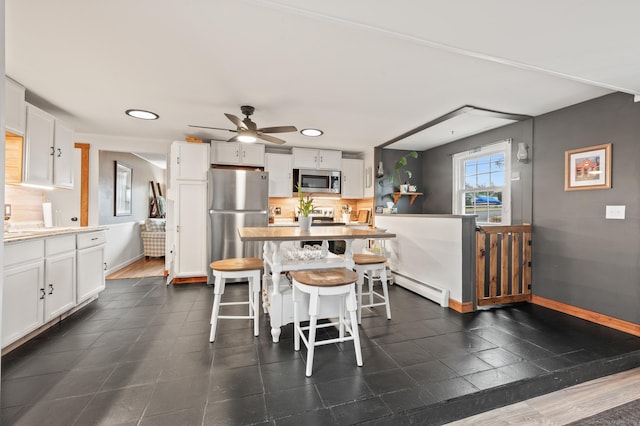 kitchen with ceiling fan, baseboard heating, a kitchen bar, white cabinetry, and stainless steel appliances