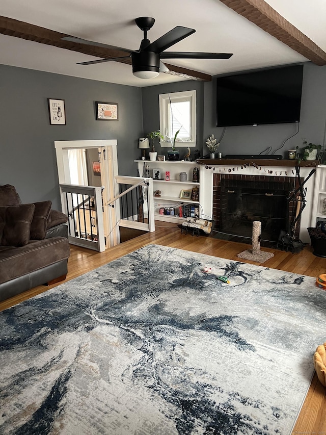 living room with ceiling fan, beamed ceiling, wood-type flooring, and a brick fireplace