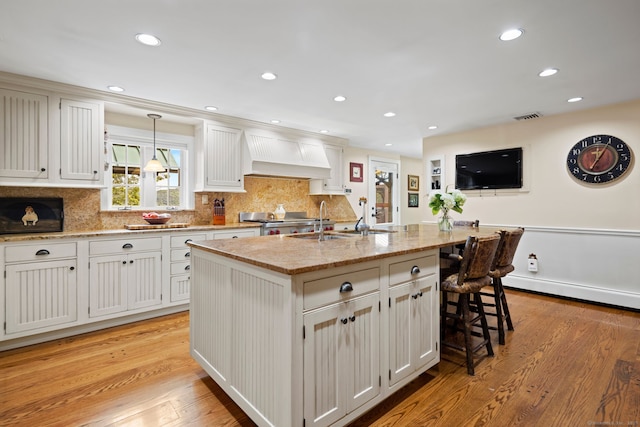 kitchen featuring white cabinets, custom range hood, light wood-type flooring, and a center island with sink