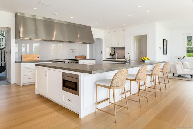 kitchen featuring dark countertops, stainless steel microwave, a large island with sink, and wall chimney range hood