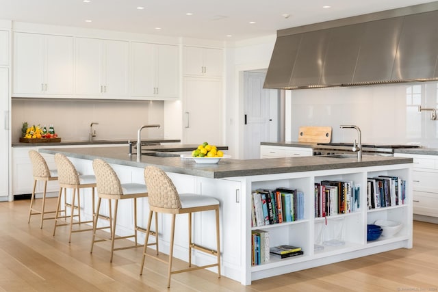 kitchen featuring a large island with sink, light wood-type flooring, white cabinetry, and wall chimney exhaust hood