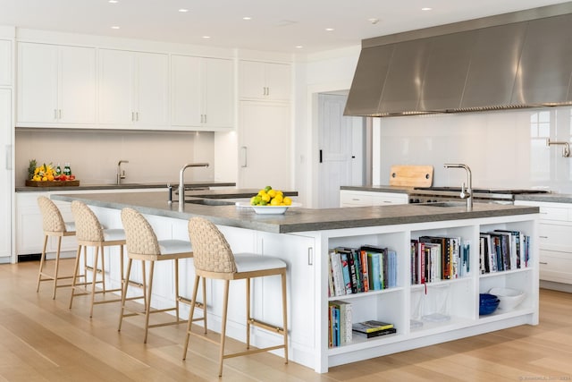 kitchen with dark countertops, light wood-style floors, white cabinets, a kitchen island with sink, and wall chimney range hood
