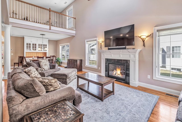 living room featuring a tile fireplace, a towering ceiling, ornate columns, and light wood-type flooring