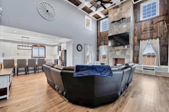 living room with coffered ceiling, a stone fireplace, beamed ceiling, a towering ceiling, and light wood-type flooring