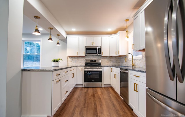 kitchen with white cabinetry, kitchen peninsula, appliances with stainless steel finishes, hanging light fixtures, and dark stone counters