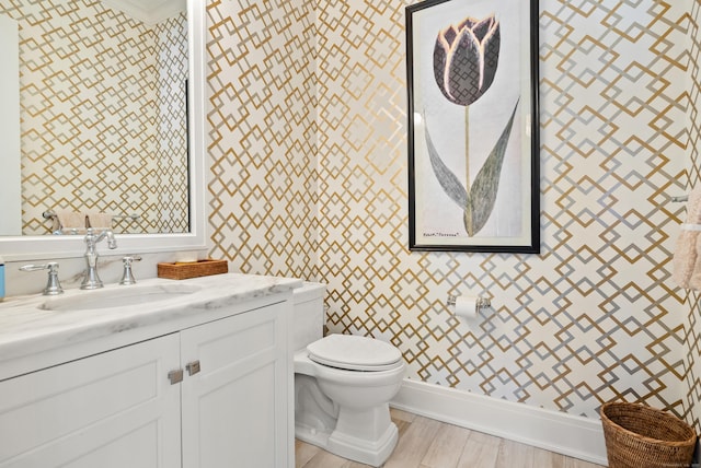 bathroom featuring toilet, vanity, wood finished floors, baseboards, and wallpapered walls