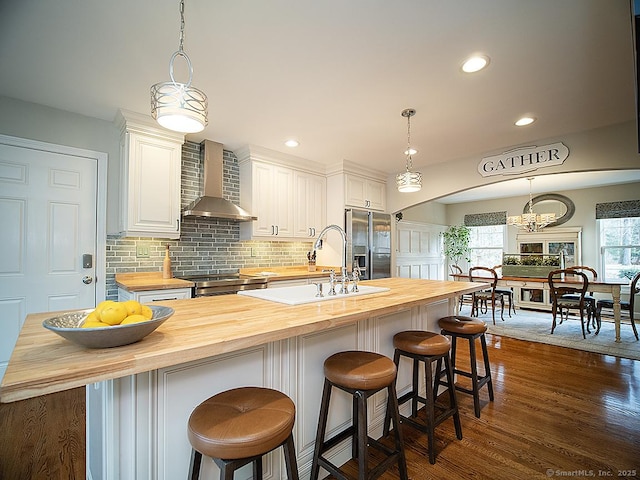 kitchen with wood counters, white cabinets, a center island with sink, wall chimney range hood, and sink