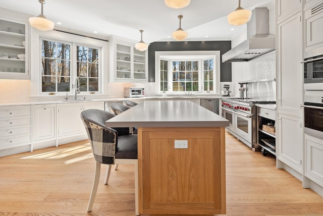 kitchen featuring stainless steel appliances, a kitchen island, wall chimney exhaust hood, white cabinets, and decorative light fixtures