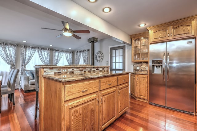 kitchen featuring decorative backsplash, stainless steel refrigerator with ice dispenser, ceiling fan, dark wood-type flooring, and a wood stove