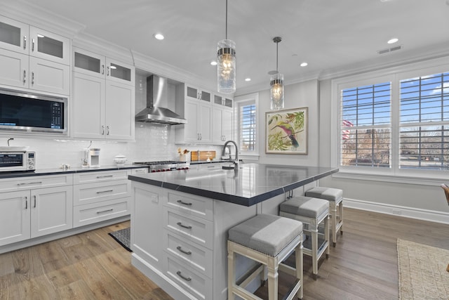 kitchen featuring a center island with sink, wall chimney exhaust hood, ornamental molding, white cabinetry, and stainless steel appliances